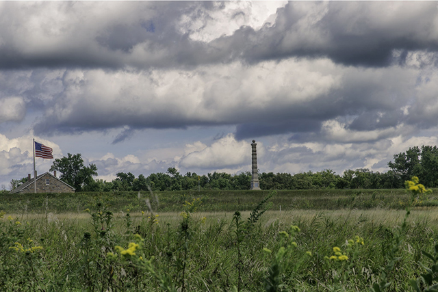 Grassy foreground with yellow flowers below a blue but cloud-filled sky, with a flag at left and a pillar at center.